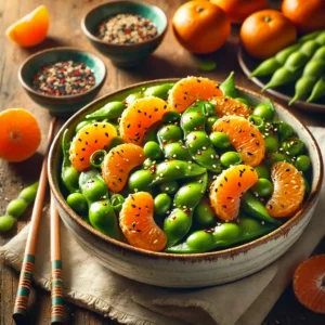 A white bowl of sweet and spicy edamame coated in glaze, topped with mandarin orange slices, sesame seeds, and green onions, set on a wooden table with fresh mandarins and chopsticks in the background.